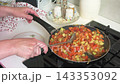 A woman prepares zucchini caviar by stirring a mix of stewed vegetables in a pan on the stove. She works in a home kitchen setting surrounded by utensils and plates. 143353092
