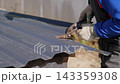 Worker in blue sleeves and gloves cuts corrugated roofing sheet with hand shears outdoors, showing precise metalwork, construction detail, and bright daylight on unfinished building site. 143359308