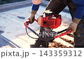Worker operates red earth auger on outdoor construction site, drilling into soil with steady hands. Close view shows gloves, workwear, metal tool, bright sunlight, and practical labor task. 143359312