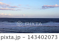Coastal waves wash toward beach under bright sky with scattered clouds. Rolling sea and distant coastline create tranquil ocean view in soft evening light, as surf advances along shore and foam 143402073