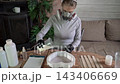 Woman wearing respirator mask and gloves pours white resin into heart-shaped silicone mold on wooden table during tabletop casting in indoor craft workshop 143406669