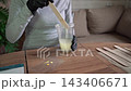 Closeup hands in black gloves stirring pale resin in clear plastic cup with wooden stick on wooden table, yellow pigment nearby in indoor craft workspace 143406671
