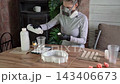 Woman in respirator and protective gloves pouring liquid into cup beside silicone mold on wooden table while mixing casting resin tools in bright home workshop 143406673