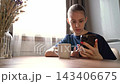 Young woman scrolling smartphone at kitchen table with white mug and dried flowers, calm indoor lifestyle moment in soft morning window light, quiet focus 143406675