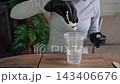 Closeup of gloved hands pouring clear liquid from small bottle into tall plastic cup above wooden table in indoor setting with plant background and soft natural light. 143406676
