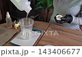 Close-up of black nitrile-gloved hands pouring clear liquid from bottle into plastic cup for resin mixing on wooden craft table in indoor workshop with safety focus 143406677
