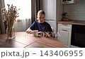 Young woman scrolling smartphone at kitchen table with coffee mug and dried flower vase, natural window daylight, calm morning lifestyle scene indoors, quiet domestic atmosphere 143406955