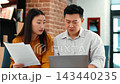 A young Asian woman and an older Asian man are sitting together at a desk. They are reviewing papers and discussing details. A laptop is open in front of them, and there are books on a shelf. 143440235