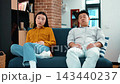 A man with dark skin and a woman of Asian descent sit on a couch in a cozy living room. They appear to be having a serious conversation about their relationship. 143440237
