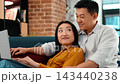 Two people are sitting on a couch, engaging in a video call. The man is showing excitement while the woman smiles at the screen. The setting includes a brick wall and cozy home decor. 143440238