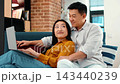 A man with dark skin and a woman of Asian descent sit side by side on a couch, focused on a laptop. They share ideas and smile while enjoying their time together in a modern living space. 143440239