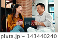 Two people are sitting on a couch in a cafe. An Asian woman smiles and talks to an Asian man, both holding coffee cups. They enjoy their time together in a warm, inviting space. 143440240