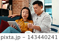 A young Asian man and a young woman with light skin are sitting on a couch at home. They are using a tablet for online shopping while the man holds a credit card. 143440241