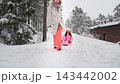A woman pulling a child on a plastic sled down a snow-covered hill in a winter forest near a traditional house during outdoor recreation. 143442002