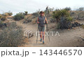 Mature woman with trekking poles walking on sandy trail enjoying an active outdoor lifestyle in dry arid landscape with desert plants and hills during her adventurous vacation. Front view in motion 143446570