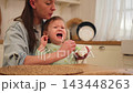 Family at home. Mother feeding her crying screaming baby girl from spoon in kitchen. Unhappy sad little toddler child with messy face expressing frustration during mealtime at home. Children emotions 143448263