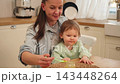Happy family at home. Mother feeding her baby girl from spoon in kitchen. Little toddler child with messy funny face eats healthy food at home. Young woman mom giving food to kid daughter 143448264