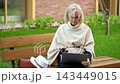 Woman writes book on laptop while seated on park bench. She keeps focused, thoughtful expression, working outdoors beside handbag and white hat in quiet green surroundings, editing digital manuscript. 143449015