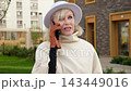 Elegant woman makes outdoor call on smartphone beside modern apartments. She wears white hat and gloves, standing in courtyard with focused expression while talking and listening on phone. 143449016