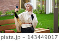 Smiling woman takes selfie on park bench with smartphone. Blonde woman in white hat poses outdoors, adjusting her look while holding phone high for self portrait in bright summer light. 143449017