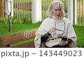 Smartphone use continues as older woman sits on wooden bench in park. She lowers her glasses, studies phone, and appears mildly concerned while resting outdoors, holding device in both hands. 143449023