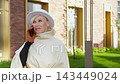 Woman with smartphone at ear smiles in outdoor phone conversation. White hatted woman stands by modern brick and glass apartments, holding steady while speaking and listening with calm confidence. 143449024