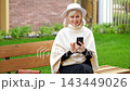 Woman on park bench checks smartphone with focused calm. Seated woman in white hat and poncho taps screen outdoors beside green grass, with tidy courtyard backdrop and relaxed expression. 143449026