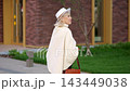 Older woman walks outdoors beside brick facade, holding brown handbag and wearing white hat. Elegant street style and relaxed expression stand out during this quiet stroll past lawn and glass. 143449038