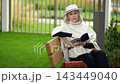 Woman reading book sits on bench in quiet green garden. She remains absorbed in pages, lifting one hand near her face as her thoughtful expression suggests peaceful pause. White hat shades her face. 143449040