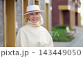 Blonde woman stands in urban portrait near modern building columns. Her white hat and cream turtleneck frame calm smile as elegant walkway and muted facade stay softly blurred behind her. 143449050