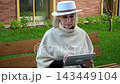 Woman on park bench uses tablet for outdoor browsing. She studies screen with thoughtful expression, then pauses with one hand near her chin in calm garden setting beside brick walls. 143449104