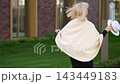 Elegant older woman poses outdoors with white hat, smiling softly in cream cape and black dress. Fashion model adjusts brim and turns beside modern building with graceful, poised confidence. 143449183
