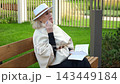 Writer on park bench holds open notebook and pen, working through notes in garden setting. Elderly woman pauses between lines, looking upward with thoughtful, composed concentration. 143449184