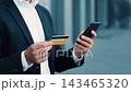 Focused Businessman Holding Credit Card And Smartphone While Making An Online Payment At Urban Setting. Professional Male In Formal Attire Engaging With Financial Transaction Outdoors. Business 143465320