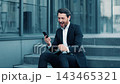 Cheerful Caucasian Man Smiling While Celebrating Success On Smartphone In Outdoor Corporate Setting, Sitting On Steps. Engaging Emotion Marginally Representative Of Business Concept. 143465321