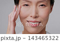 Facial Skincare. Happy Mature Chinese Lady Applying Moisturizer Cream On Cheek Moisturizing Face Caring For Skin Posing Over Gray Studio Background, Smiling To Camera. Slowmo 143465322
