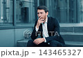 Worried Man In Formal Suit With Short Beard, Sitting On Steps Outside, Covering Face With Hand, Expressing Stress At Workplace, Outdoor Business Setting. Business Concept. 143465327