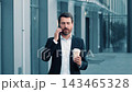 Focused Male Businessman Holding Coffee Cup And Talking On Phone While Walking In Urban Setting, Demonstrating Professional Lifestyle And Business Concept. Engaged And Productive Moment. 143465328