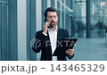 Focused Caucasian Male Businessman Speaking On Smartphone While Holding Clipboard In Outdoor Office Setting, Representing Business Concept And Professionalism In Corporate Environment. 143465329