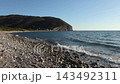 Pebble shoreline, gentle waves lapping against the stones, a tree-covered headland in the distance, and the afternoon sun 143492311