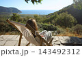 A young woman relaxes in a wooden deck chair, on the horizon lie coastal hills and the sea, bathed in the golden light of the afternoon sun. 143492312