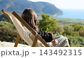 Woman relaxing on wooden deck chair as black cat curls on lap, overlooking coastal hills, sea horizon, golden afternoon sunlight over lush greenery serene outdoor lifestyle moment 143492315