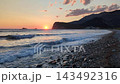 Golden sunset over pebble beach with gentle waves lapping shoreline, silhouetted headland, and scattered clouds in warm sky 143492316