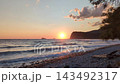 Golden sunset over pebble beach with gentle waves lapping shoreline, silhouetted headland, and scattered clouds in warm orange sky 143492317