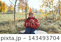 Portrait of caucasian child who smiles and looks at camera with pumpkin in hands in autumn park. Boy in anticipation of holiday on sunny day sits on rock. 143493704
