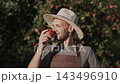 Caucasian man in traditional hat stands in apple orchard and holds ripe apple and smiles as he inhales its fragrance. Cultural heritage meets nature in warm sunlight. 143496910