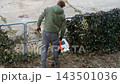 A man uses a gasoline leaf blower to clear yard debris along a metal fence, demonstrating efficient garden maintenance and property cleanup tasks in a residential spring setting 143501036
