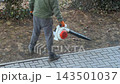 A worker using a handheld power leaf blower to clear yard debris along a paved walkway, demonstrating efficient garden maintenance and lawn care services in a residential outdoor space 143501037