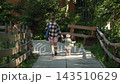 A little girl pink dress carries bundle sticks, holding mother hand look concentration, shot behind showing family strolling along paved path leading down through woods toward wooden houses warm 143510629