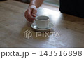 A hand lifts a white cup from a wooden table; the saucer is empty. Live-action 4K. 143516898